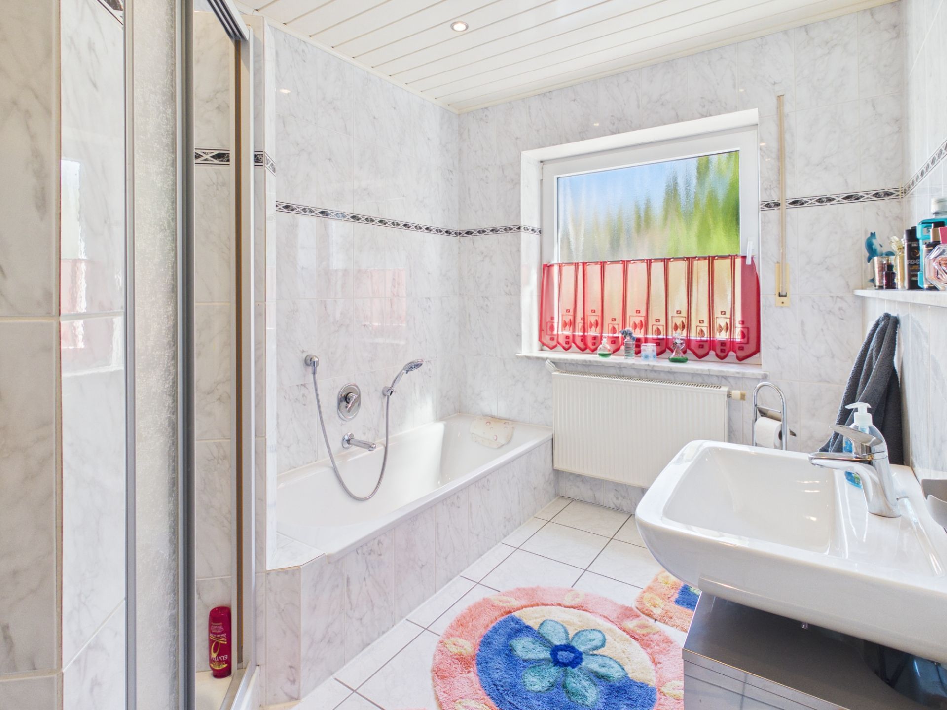Helles Bad mit Badewanne, Dusche, Waschbecken und Fenster mit rotem Vorhang.