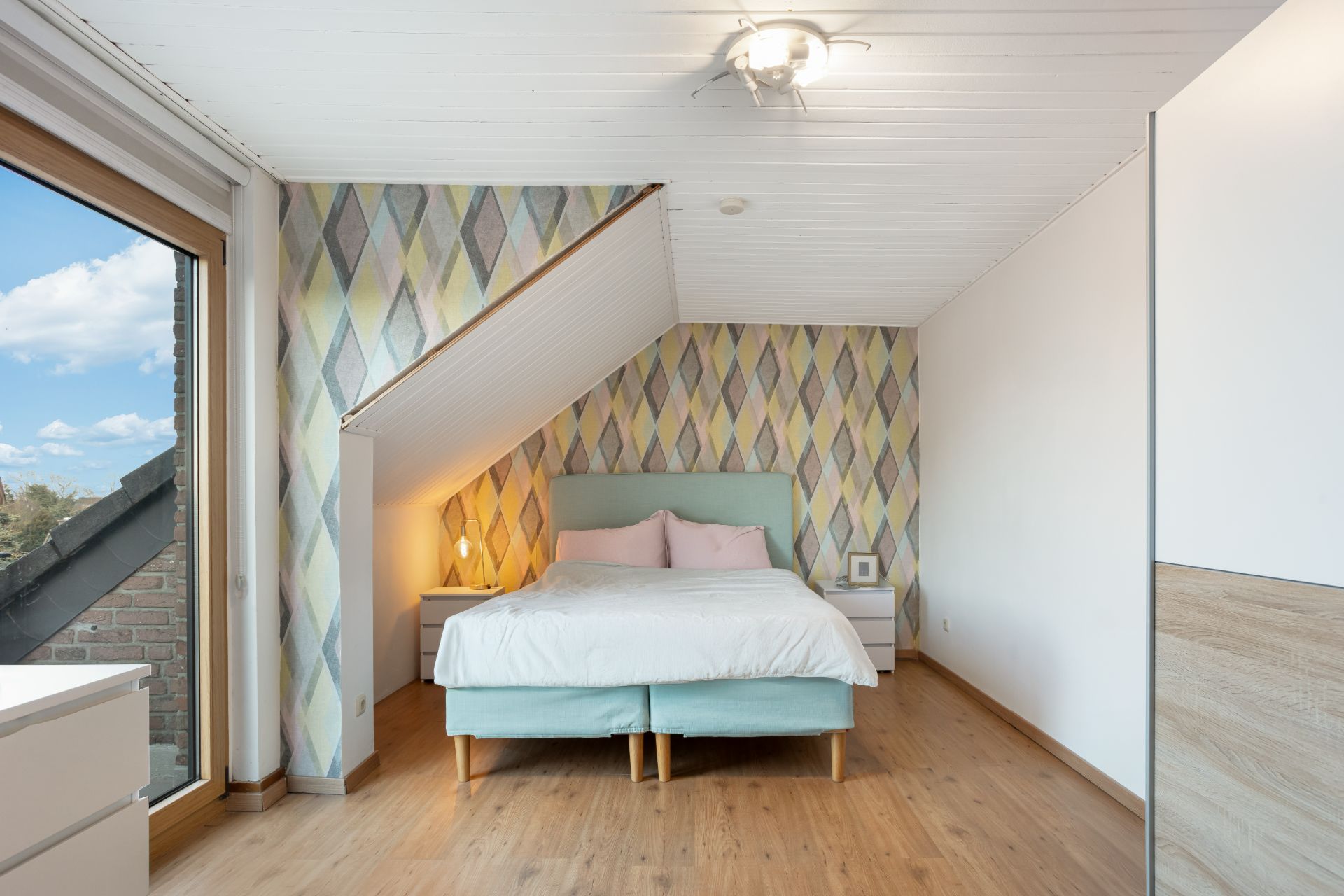 Schlafzimmer mit Doppelbett, bunter Tapete und großem Fenster auf der linken Seite.