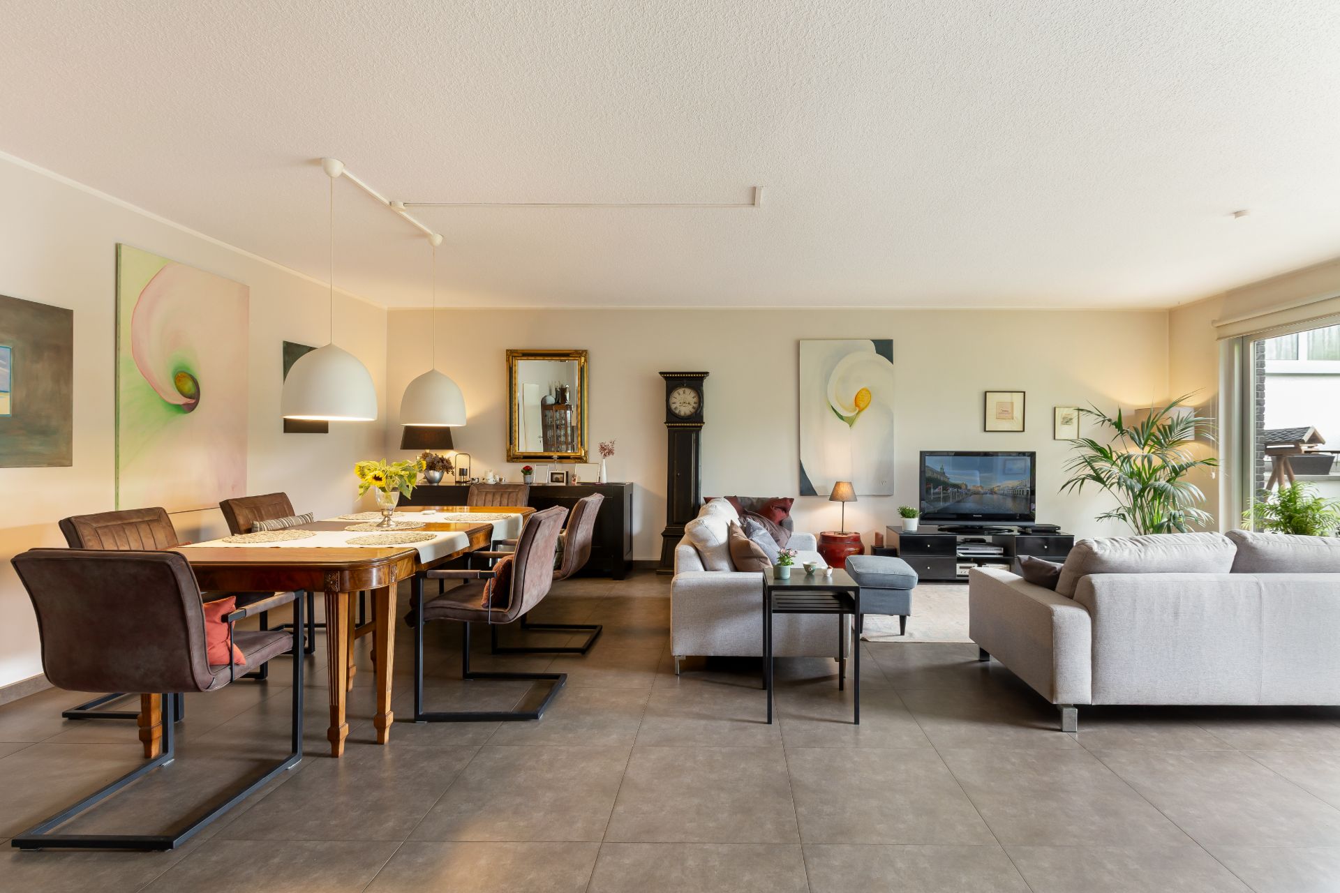 Helles Wohnzimmer mit Esstisch, Sofas, Fernseher, Standuhr und großen Fenstern.