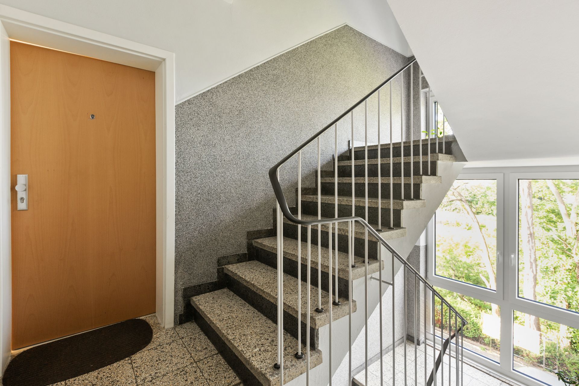 Treppenhaus mit Metallgeländer, Fenster und einer Wohnungstür links.