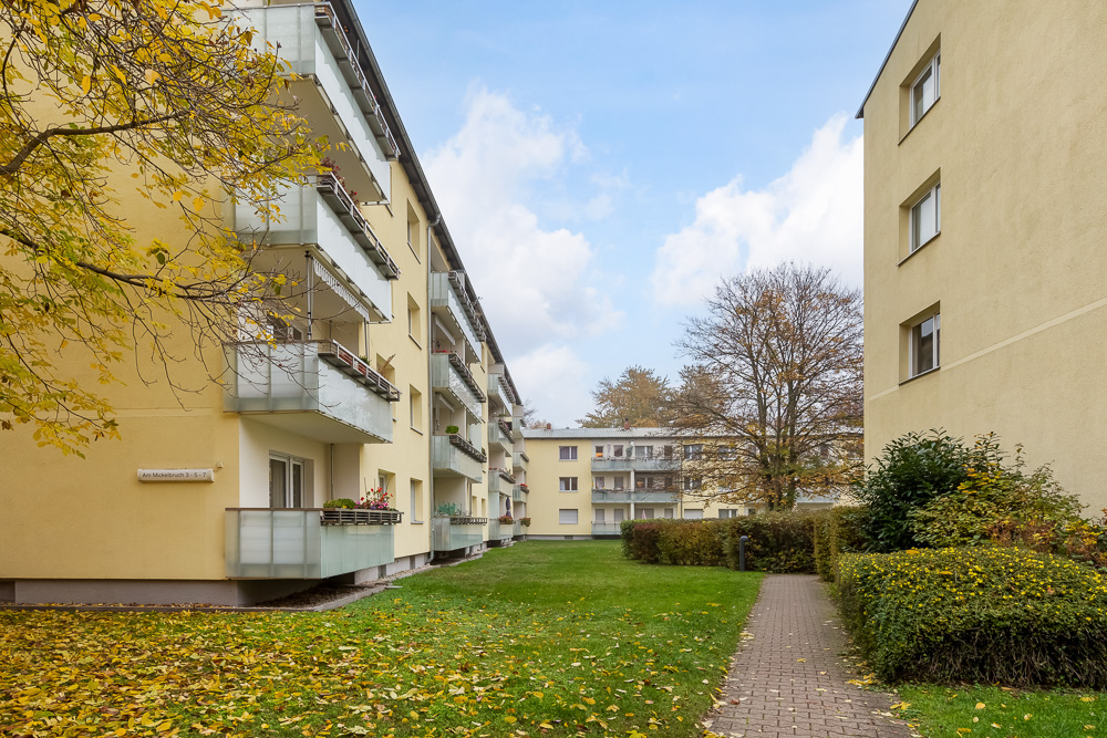 Mehrfamilienhäuser mit Balkonen und gepflegtem Rasen, Gehweg entlang der Hecke, herbstliche Bäume.