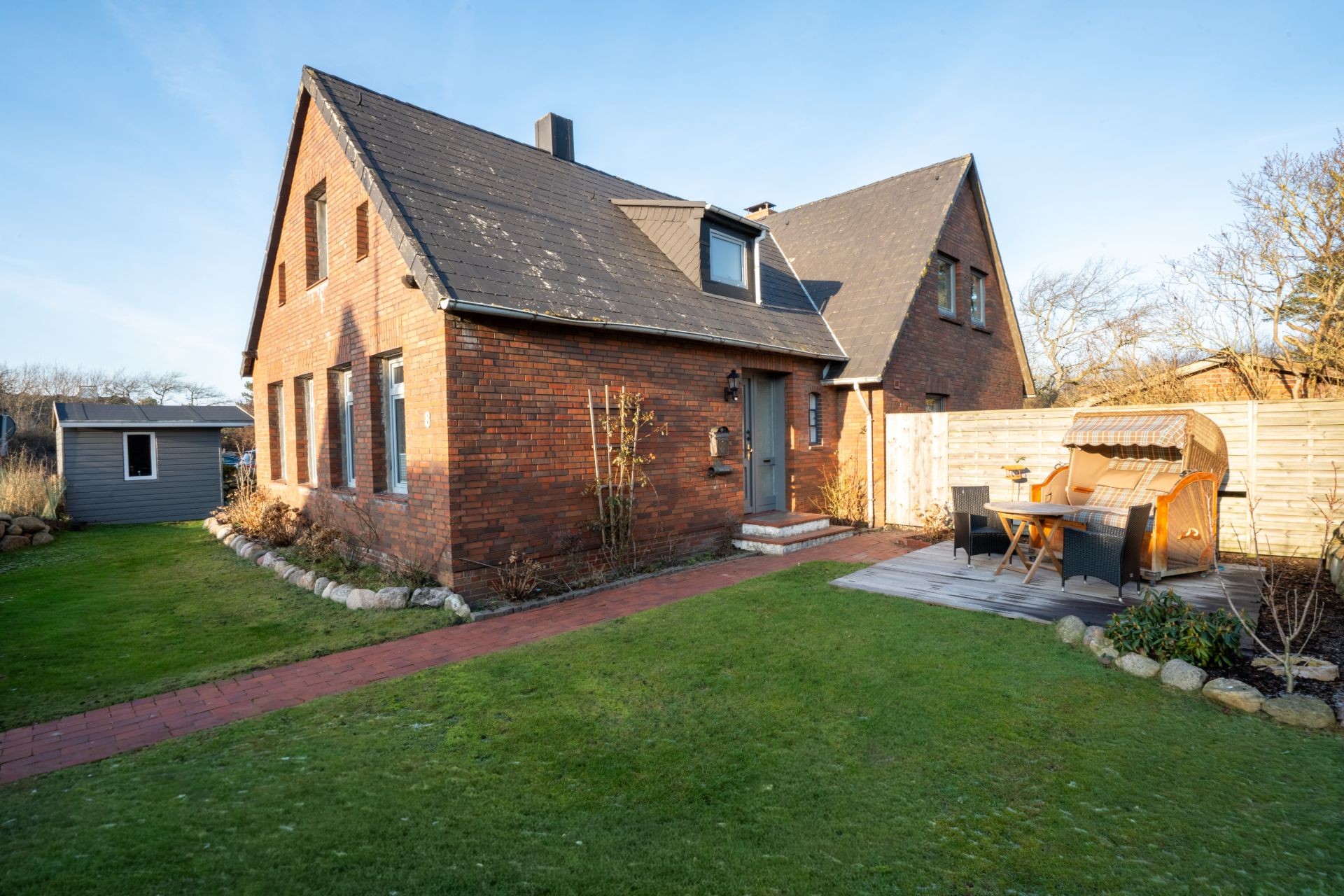 Backstein-Einfamilienhaus mit Garten, Terrasse und Strandkorb bei sonnigem Wetter.