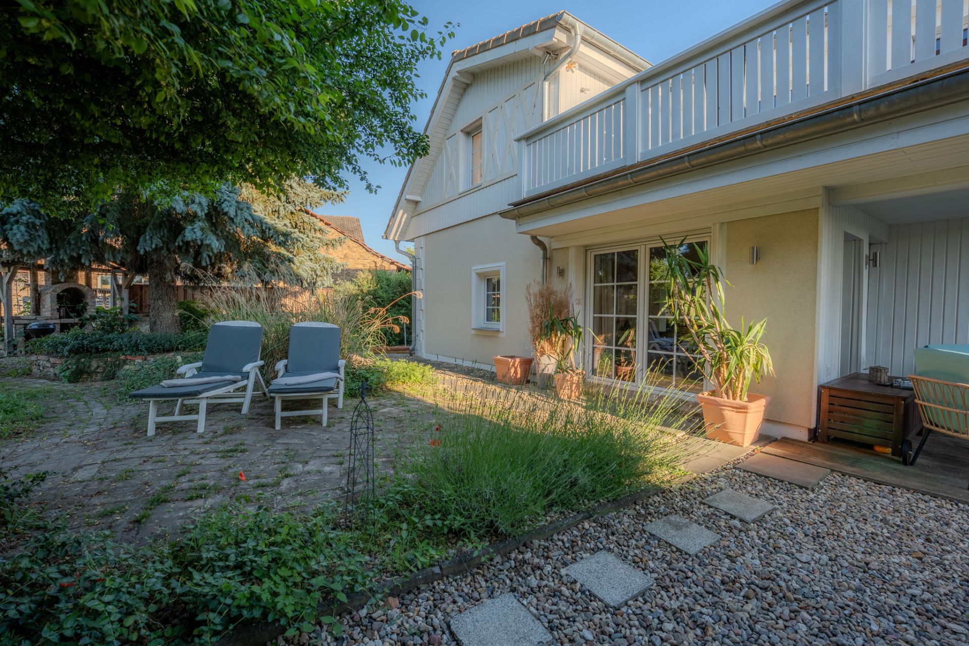 Garten mit zwei Liegestühlen vor einem hellen Wohnhaus mit Terrasse und Pflanzen.