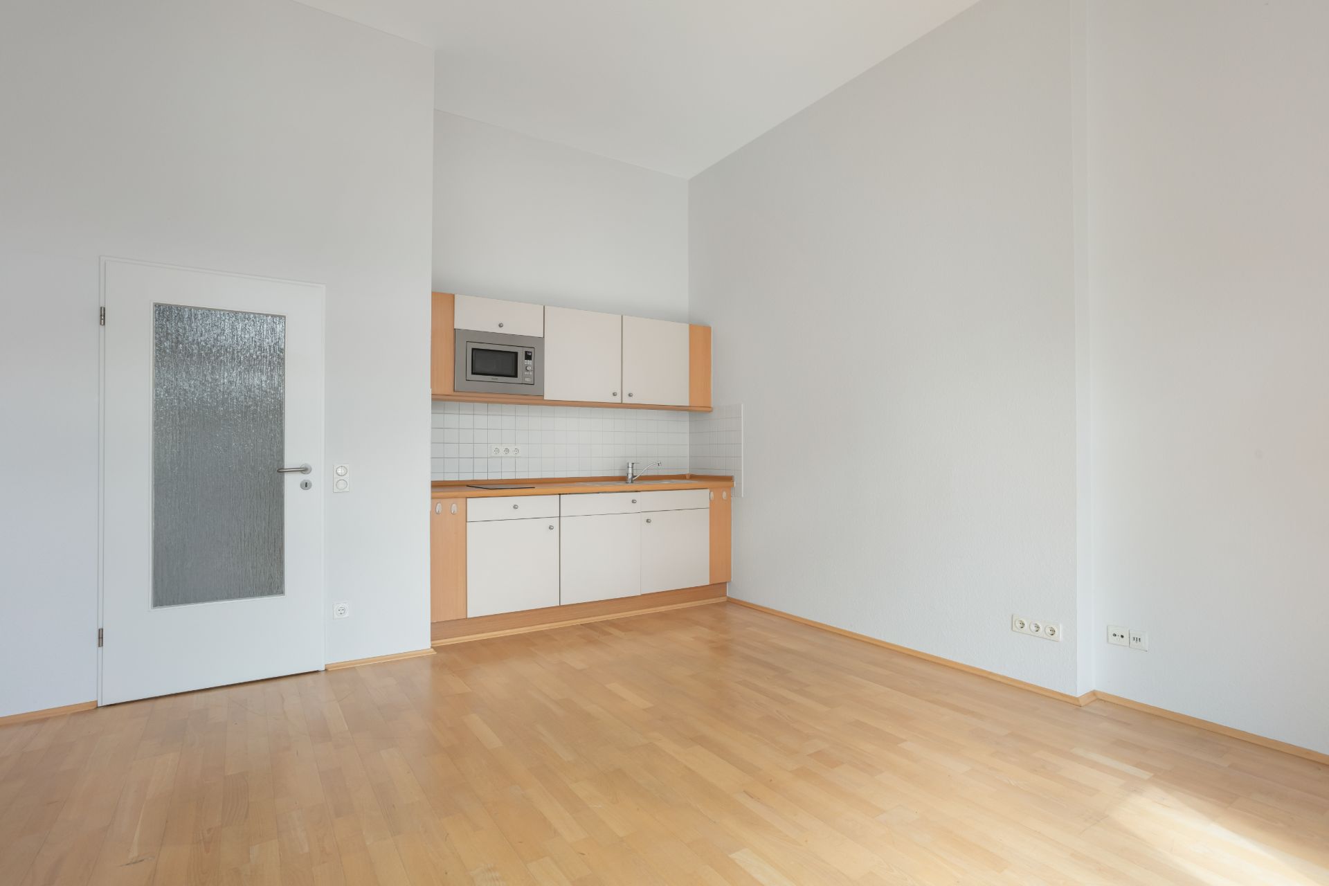 Leerer Raum mit Einbauküche, Holzboden und weißer Tür auf der linken Seite.