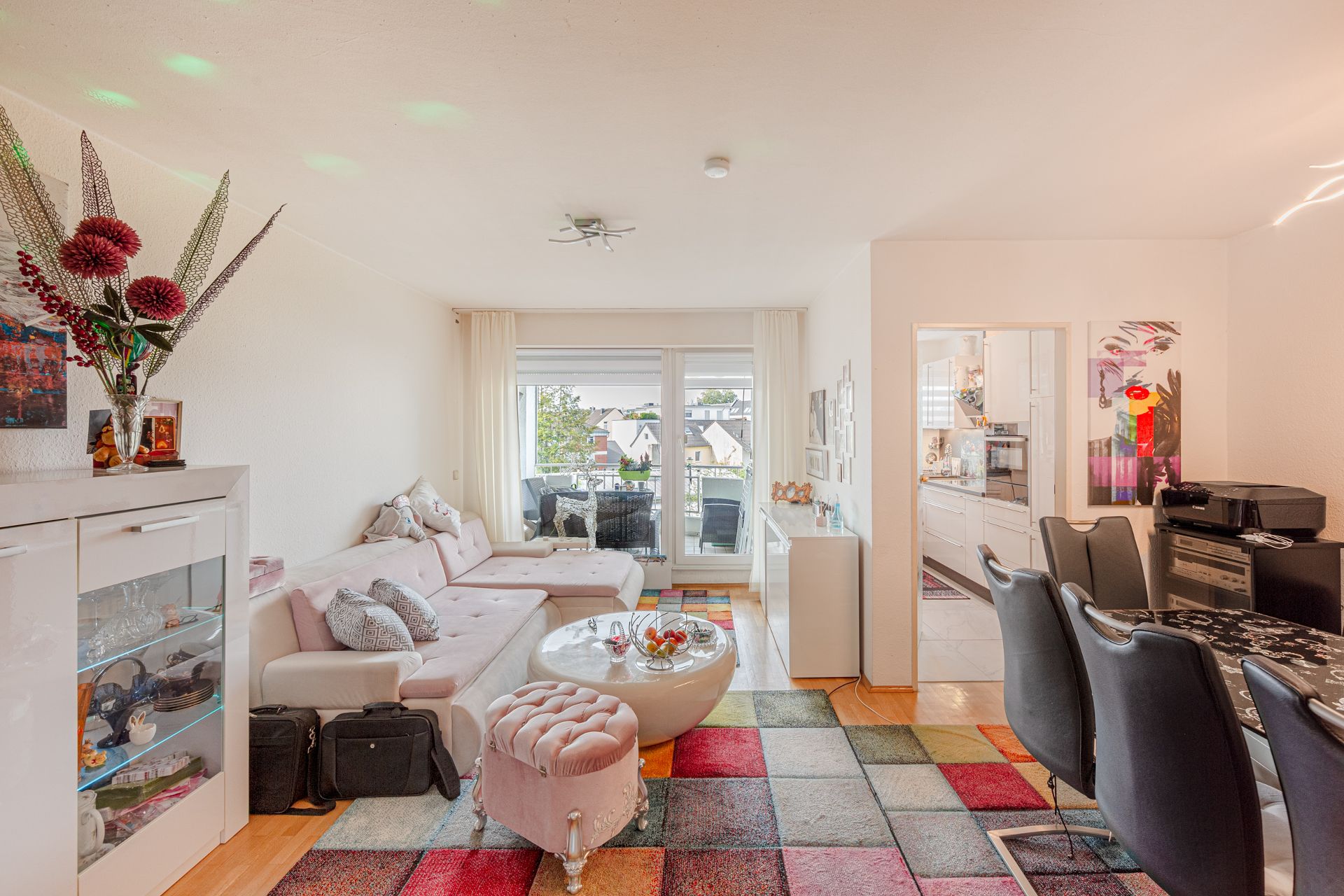 Helles Wohnzimmer mit Sofa, rundem Tisch, buntem Teppich und Essbereich, daneben Tür zur Küche.