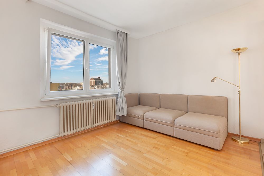 Helles Wohnzimmer mit Ecksofa, großem Fenster, Vorhang, Parkettboden und zwei Stehlampen.