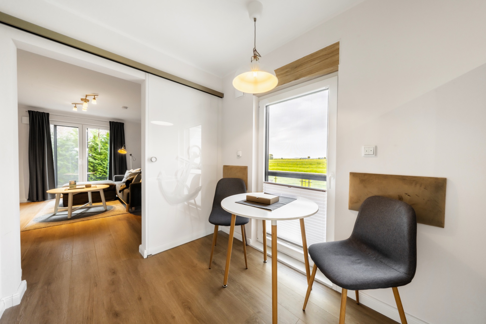 Heller Essbereich mit rundem Tisch, zwei Stühlen und großem Fenster, angrenzend an ein Wohnzimmer.