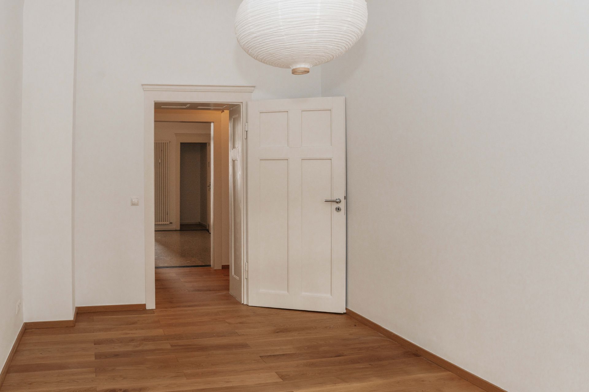 Leerer Raum mit Holzboden, weißer Wand, offener Tür und runder Lampe an der Decke.