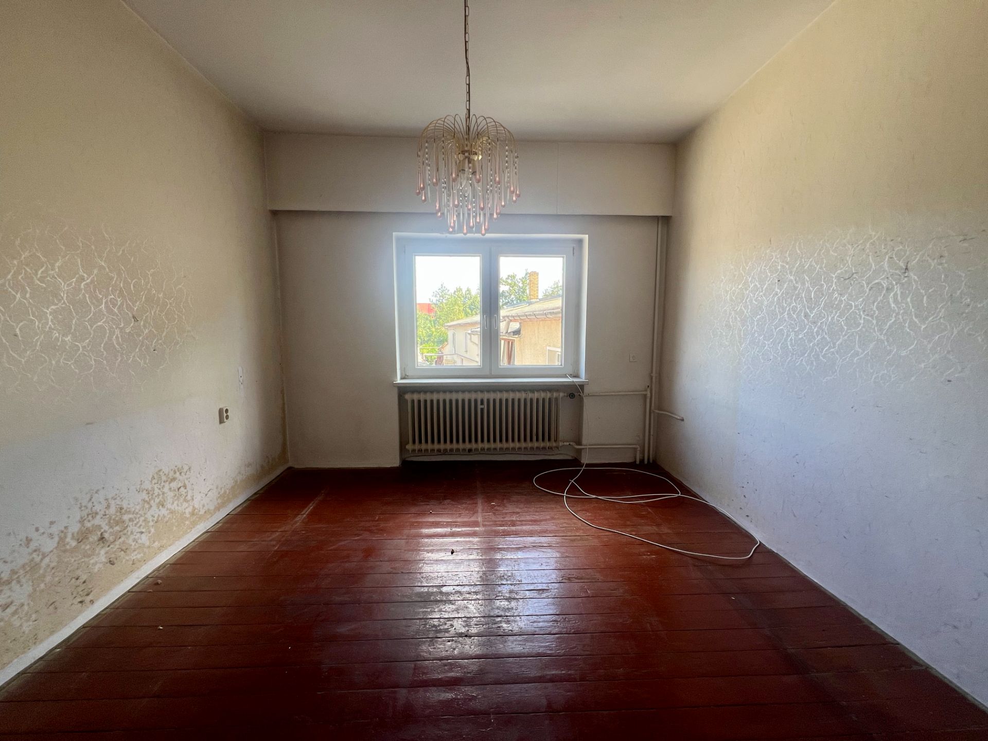 Leeres Zimmer mit abgenutztem Holzboden, Fenster, Heizkörper und Kronleuchter.