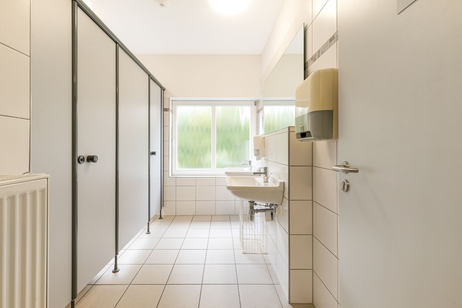 Helles Badezimmer mit zwei Waschtischen, Spiegel, Fenster und mehreren Toilettenkabinen.