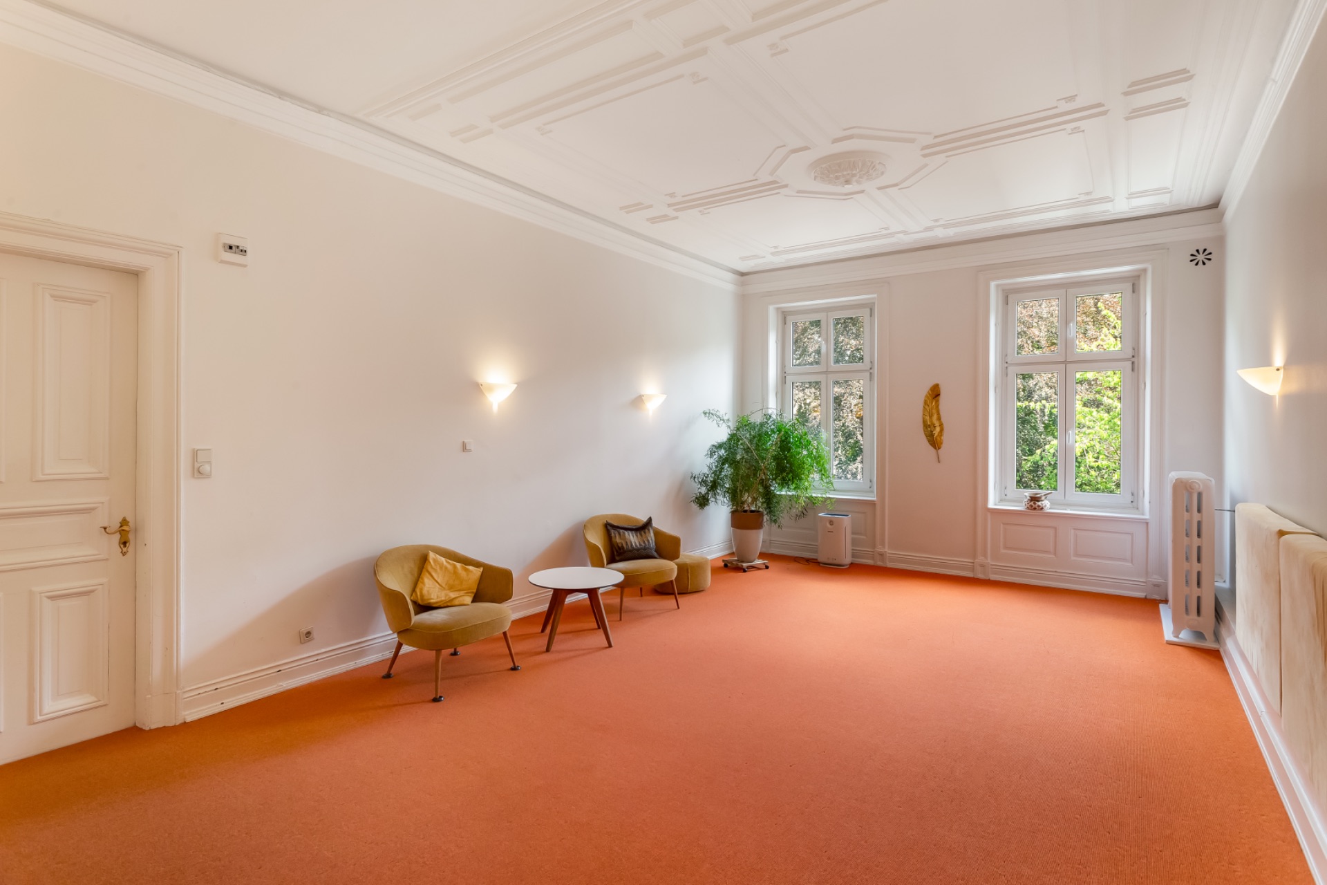 Helles Wohnzimmer mit orangefarbenem Teppich, zwei Sesseln, einem Tisch und großen Fenstern.