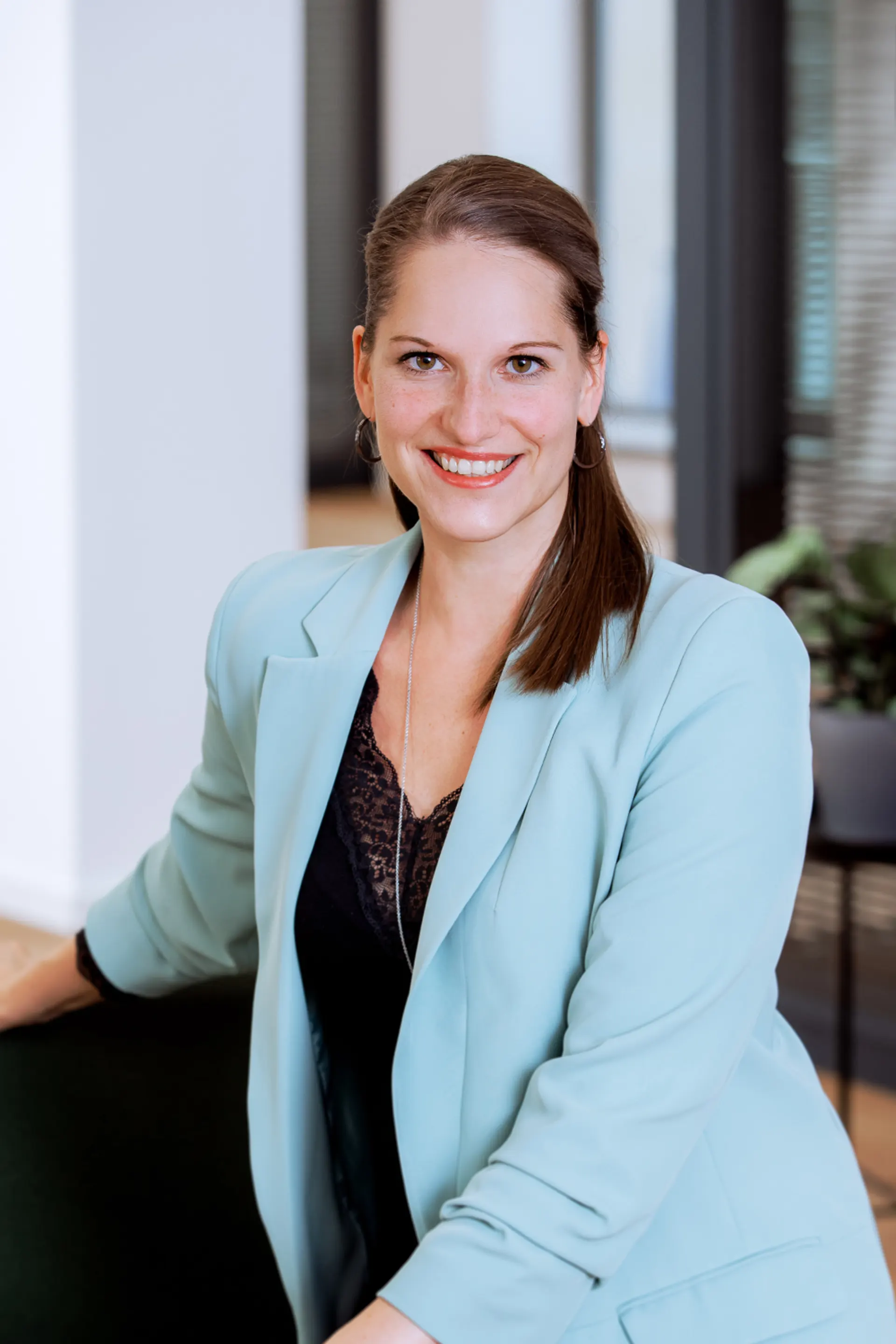 Frau mit hellblauem Blazer sitzt und lächelt in einem modernen Raum.