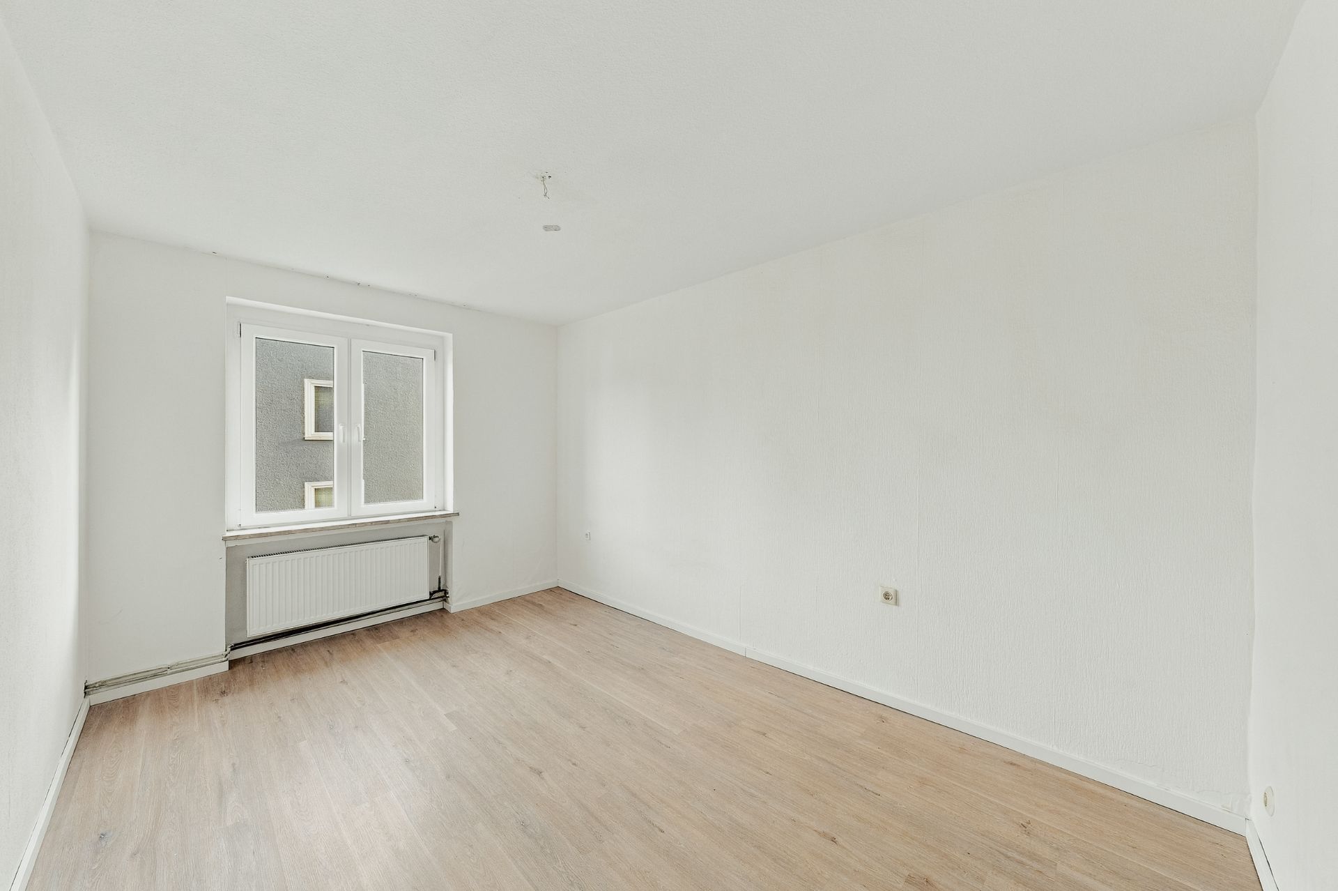 Leerer Raum mit weiß gestrichenen Wänden, Holzboden und Fenster mit Heizkörper.