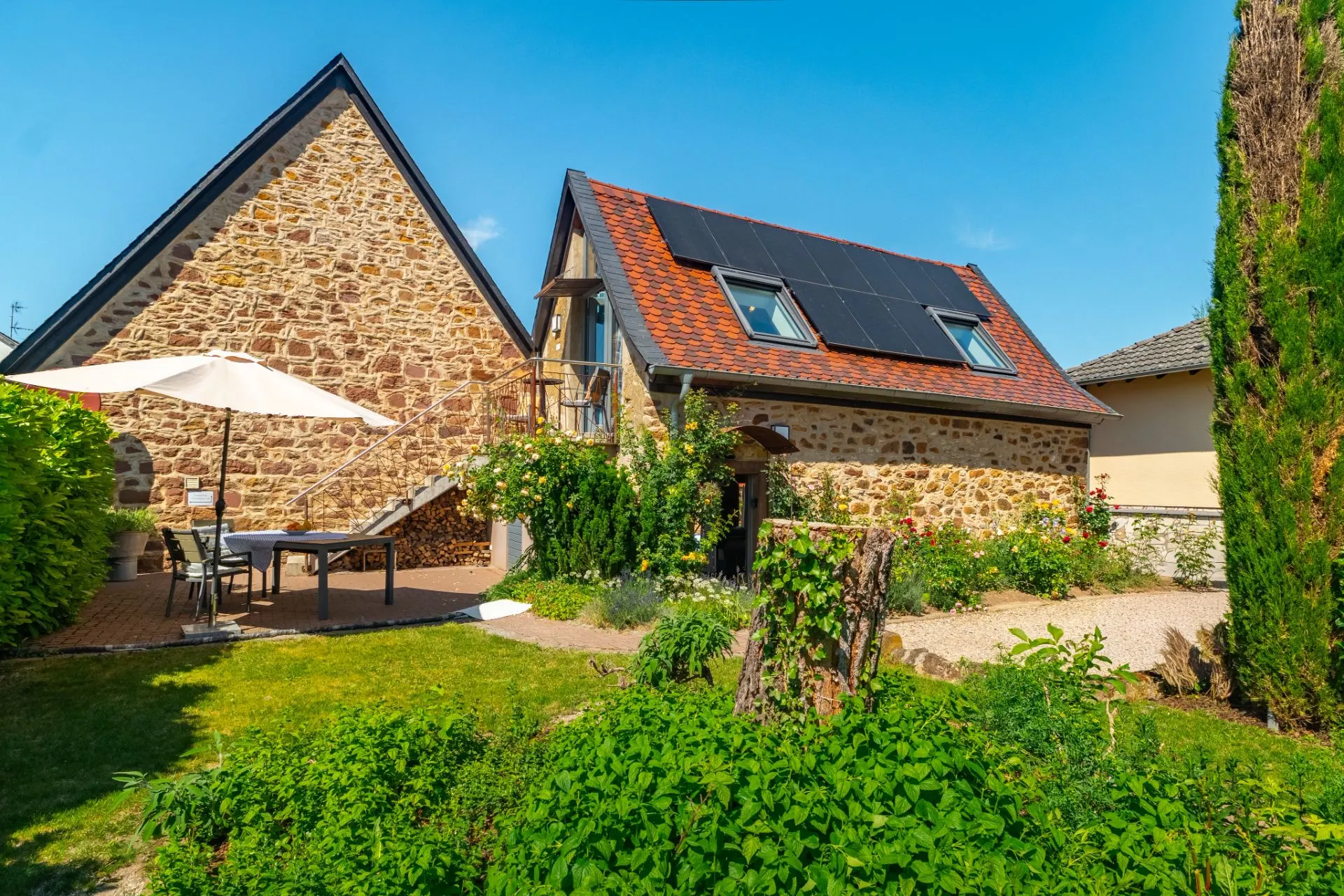 Steinhaus mit Solarpanelen, Garten, Terrasse mit Tisch und Sonnenschirm bei sonnigem Wetter.