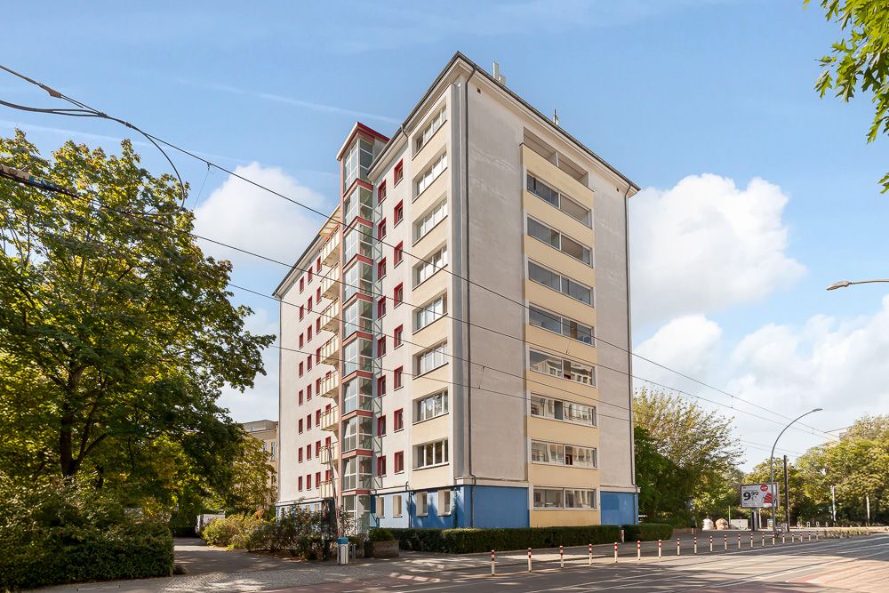 Mehrstöckiges Wohngebäude mit Balkonen an einer Straßenecke, umgeben von Bäumen.