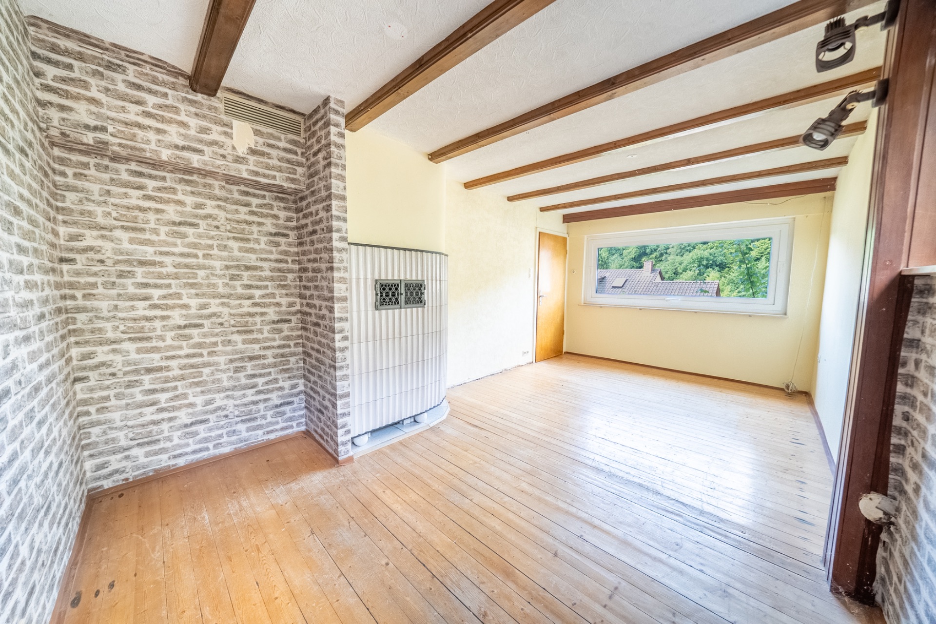 Helles Zimmer mit Holzboden, Holzbalken, Backsteinwänden und großem Fenster mit Blick ins Grüne.