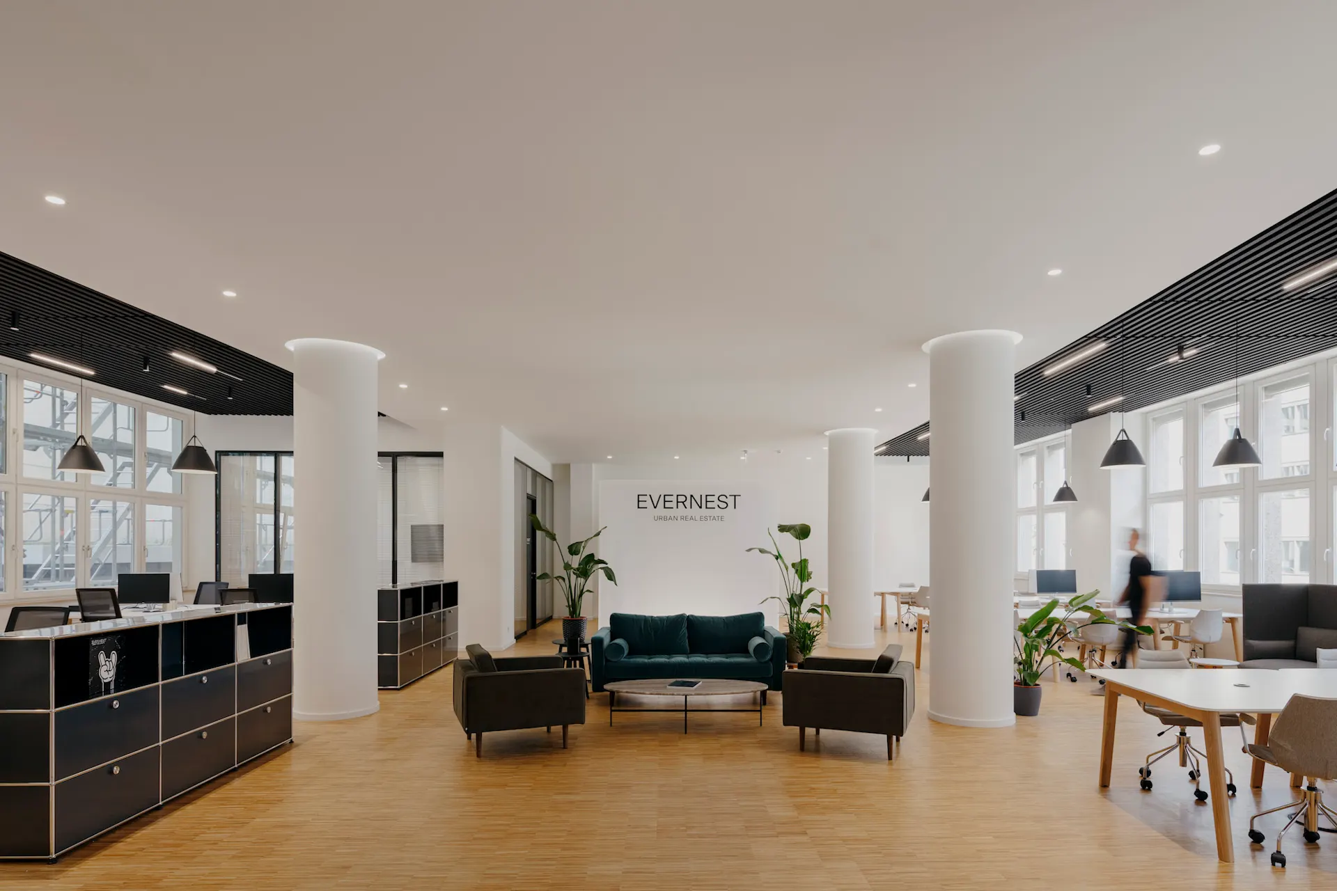 Helles Büro mit Schreibtischen, Sesseln, Sofa, Pflanzen und großen Fenstern.