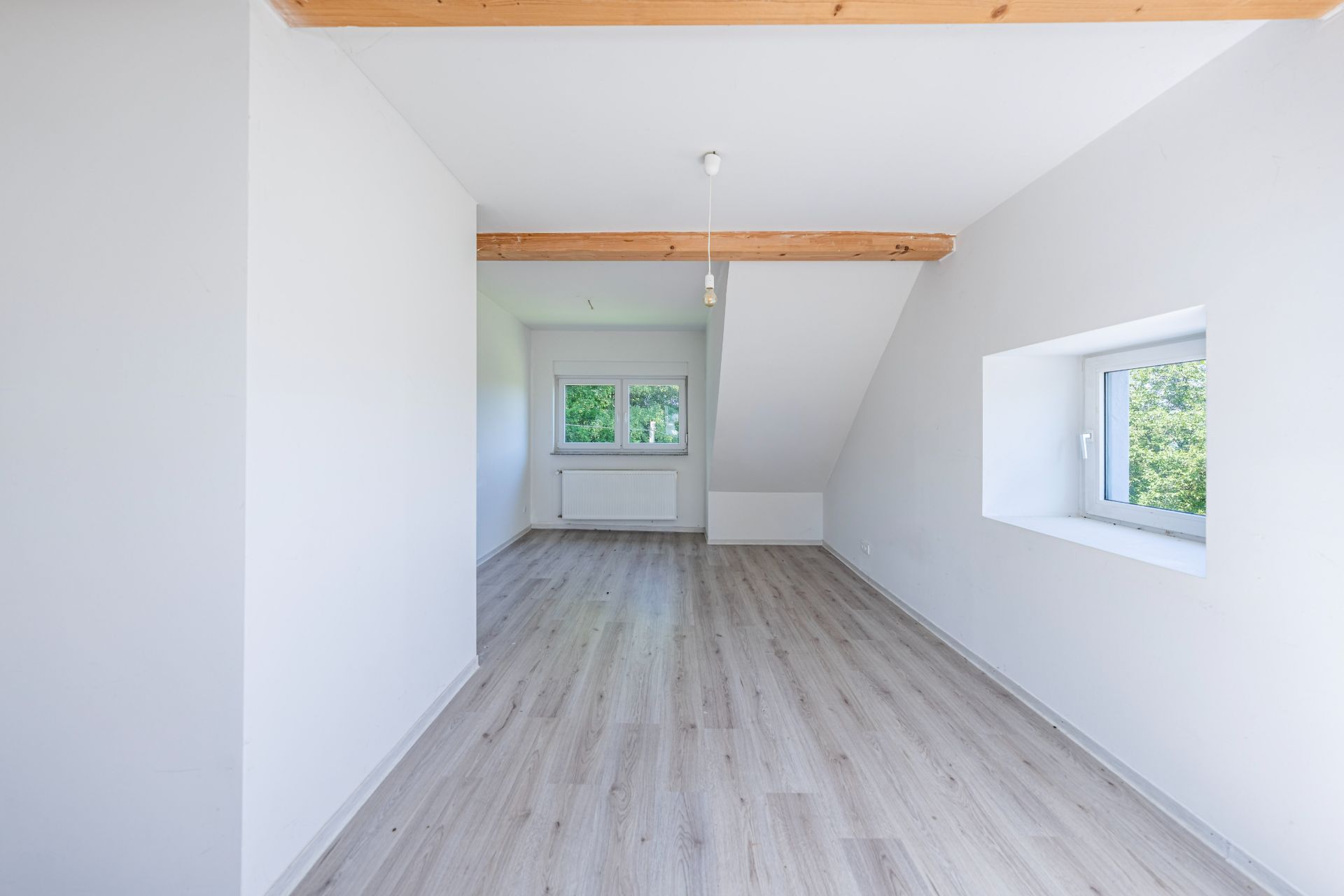 Helles, leeres Zimmer mit Holzbalken, zwei Fenstern und hellem Laminatboden.