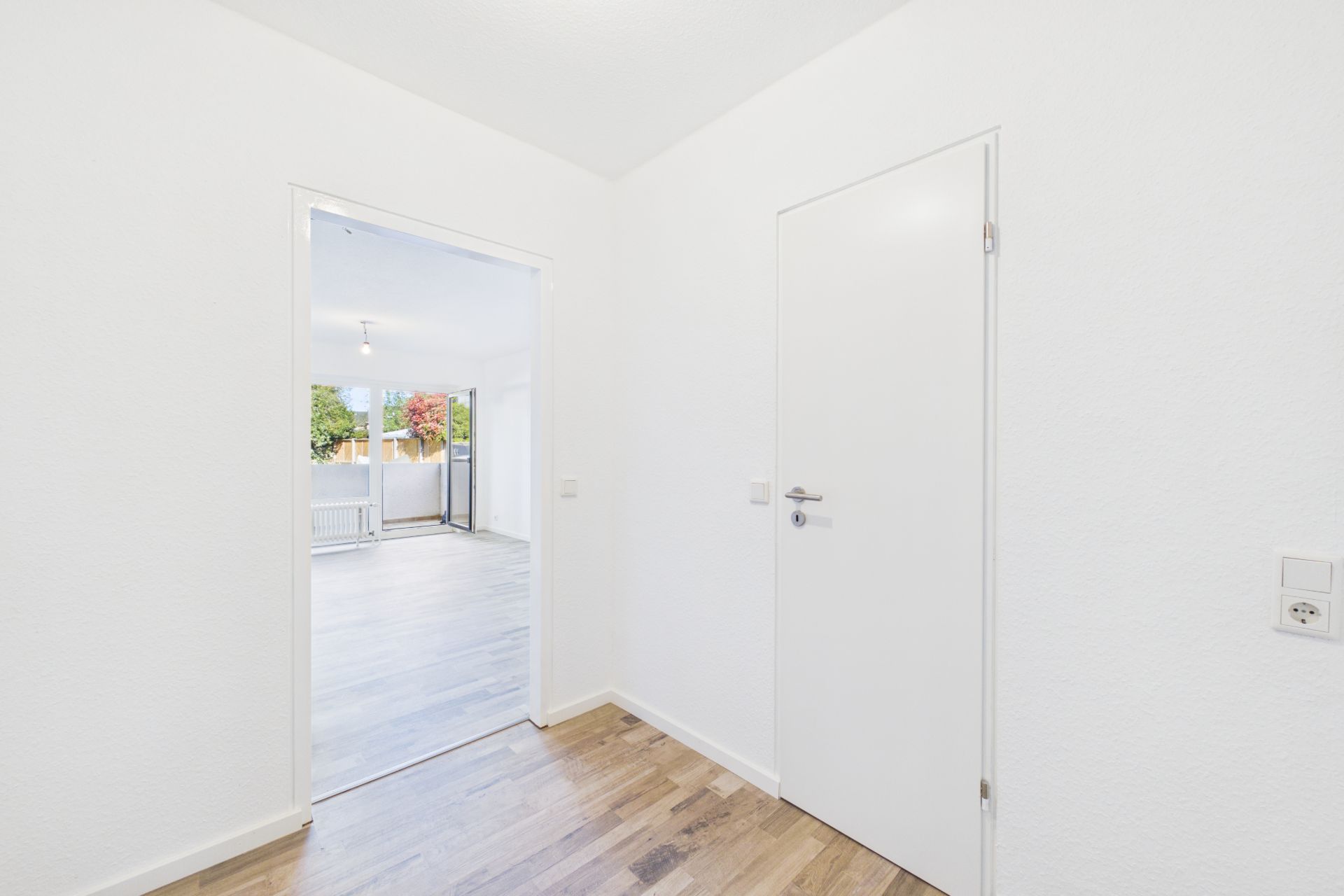 Heller Flur mit Holzboden, eine weiße Tür und offener Blick in ein Wohnzimmer mit Fenster.