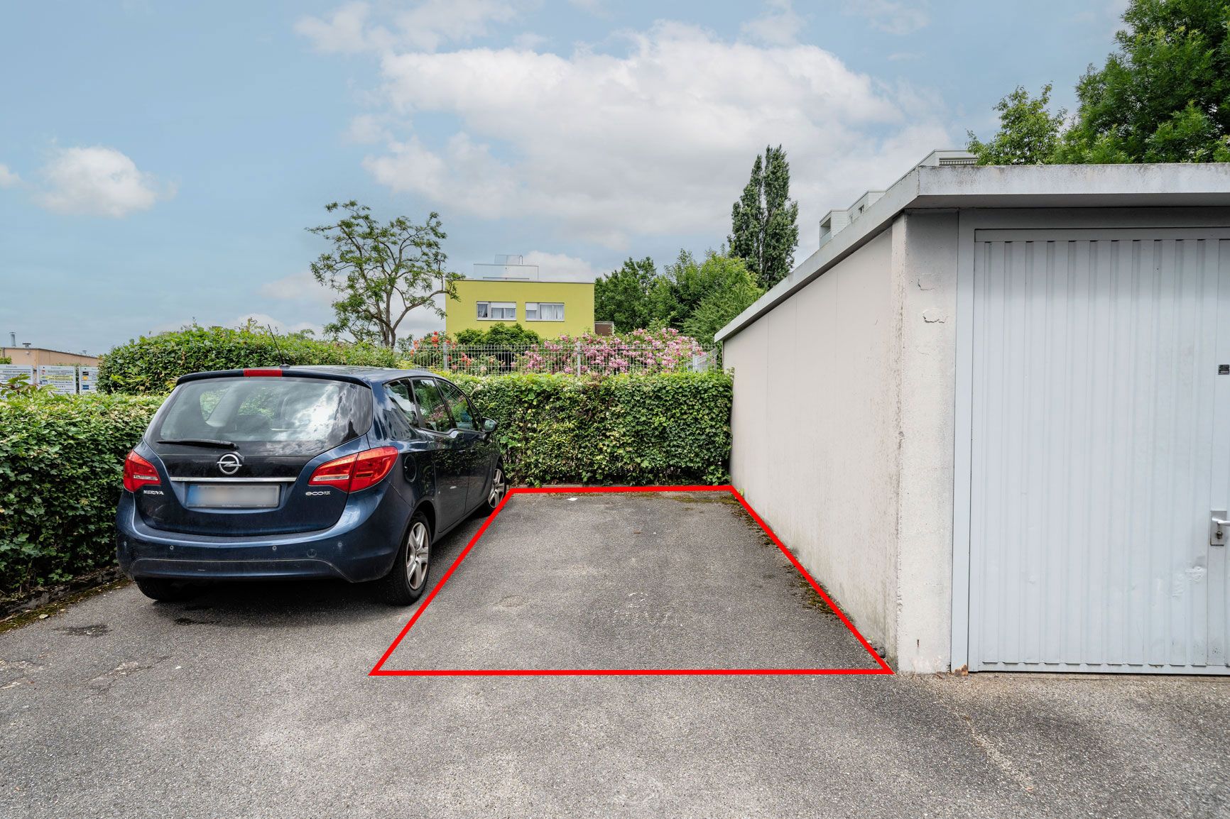 Parkplatz neben einem blauen Auto und einer weißen Garage, mit rotem Rand markiert.
