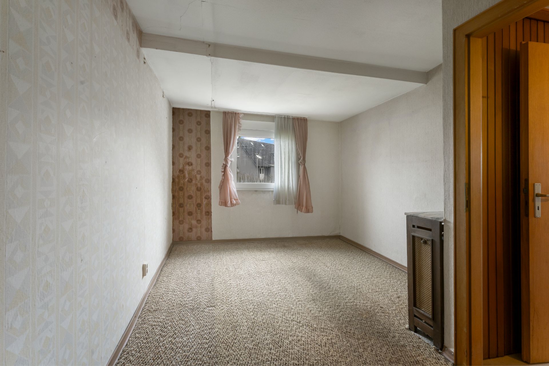 Leeres Zimmer mit Teppichboden, Fenster mit rosa Vorhängen und teilweise geöffneter Holztür.