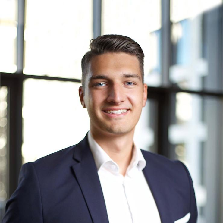 Junger Mann im Anzug steht vor großen Fenstern in einem hellen Raum.