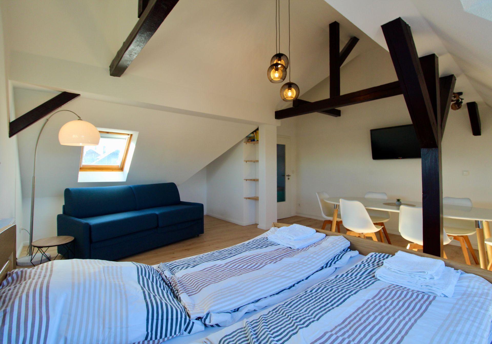 Helles Schlafzimmer mit Doppelbett, Sofa, Esstisch, TV und sichtbaren Holzbalken unter dem Dach.