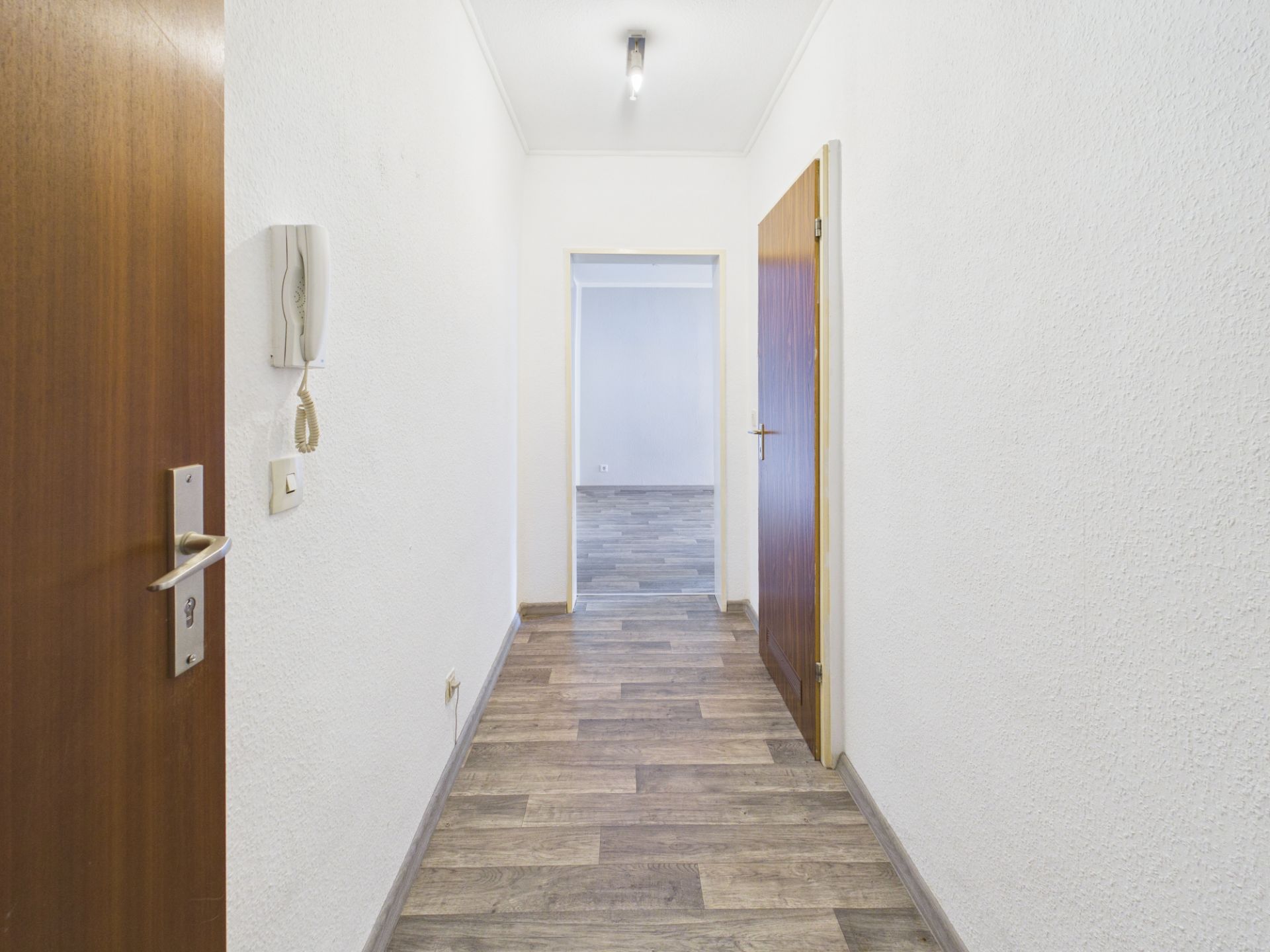 Langer, heller Flur mit Holzboden, weißen Wänden und offener Tür am Ende.