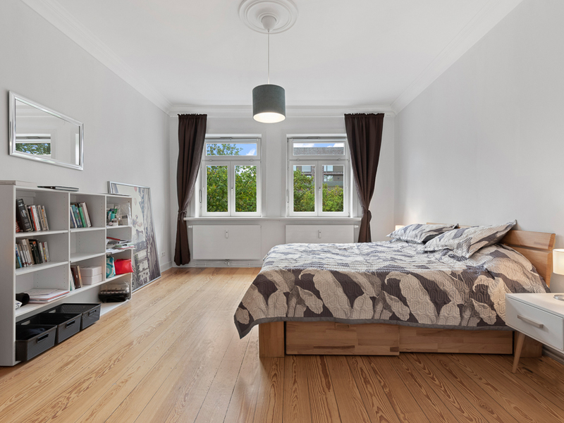 Helles Schlafzimmer mit Doppelbett, Bücherregal, Holzboden und drei Fenstern mit dunklen Vorhängen.