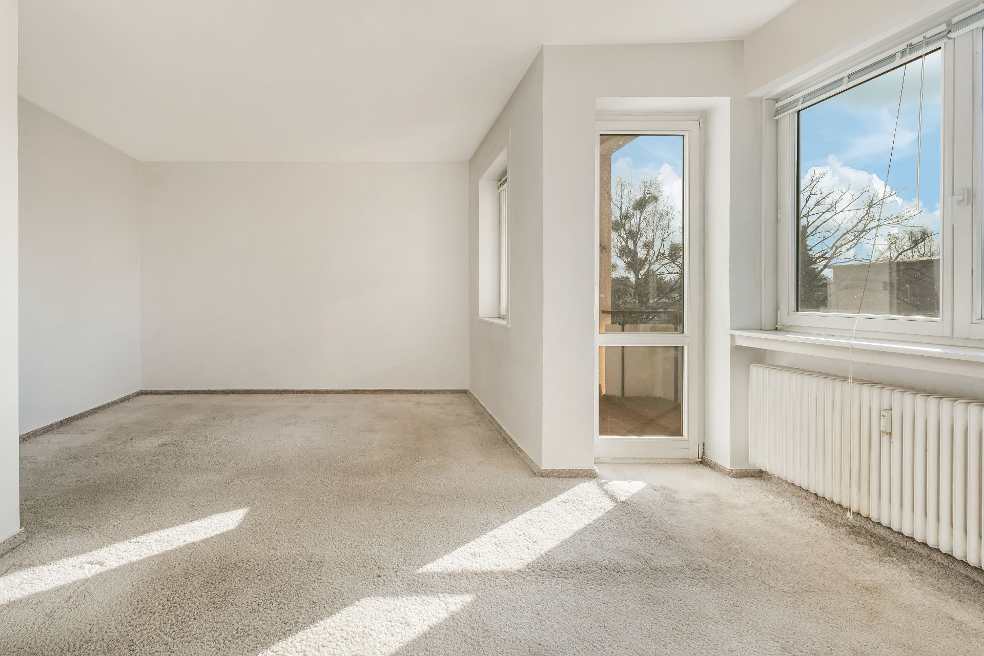 Helles, leeres Zimmer mit Teppich, großen Fenstern und Tür zum Balkon.