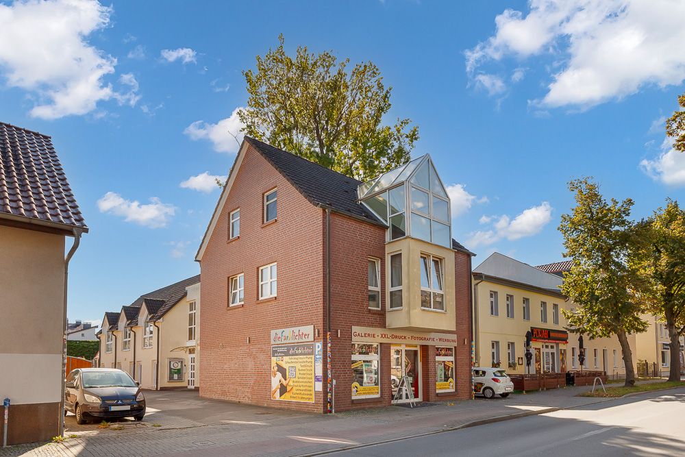 Ecke eines mehrstöckigen Wohnhauses mit Glasvorbau an einer Straße, Bäume und Autos sichtbar.