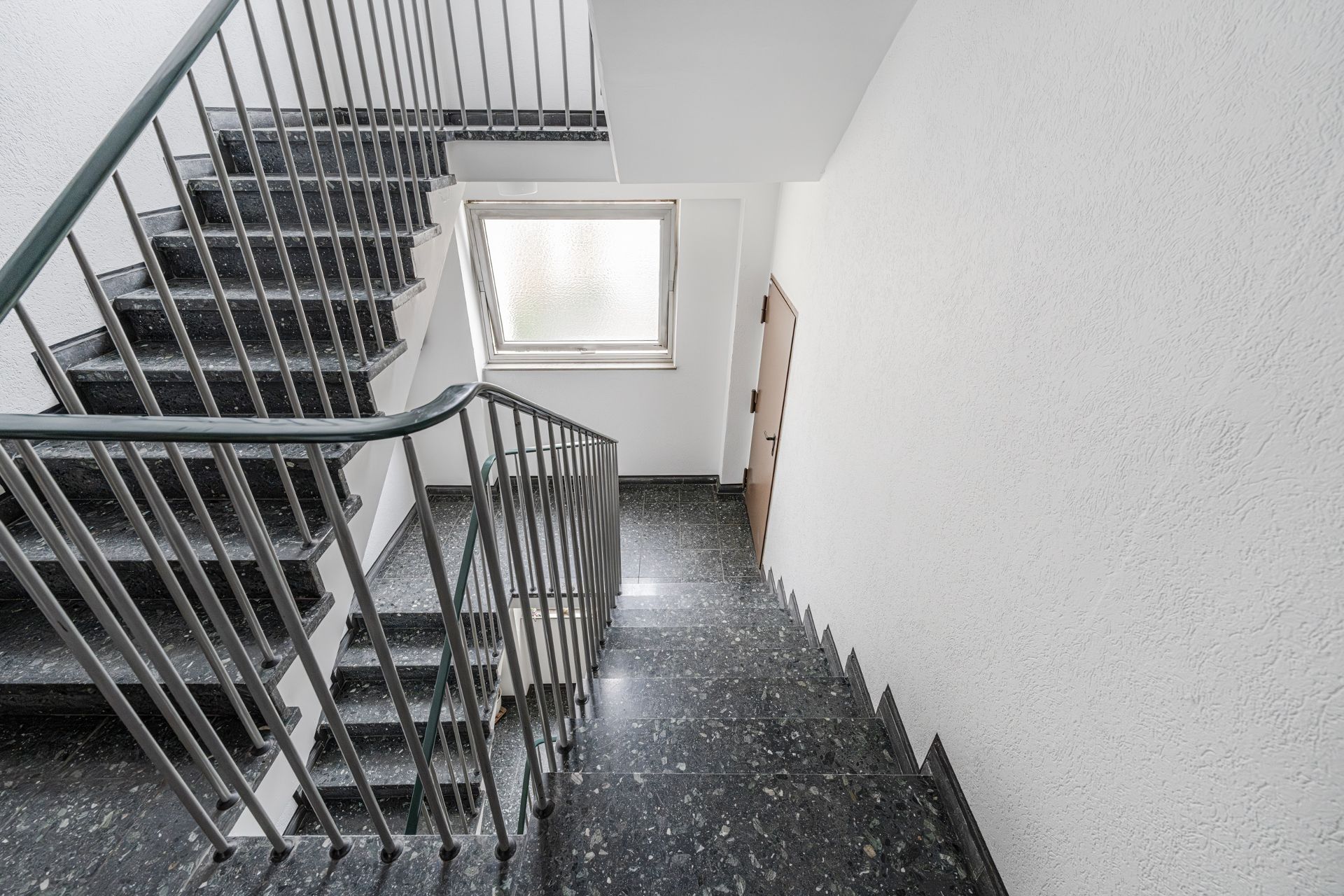 Helles Treppenhaus mit dunklen Stufen, Metallgeländer und einem Fenster.