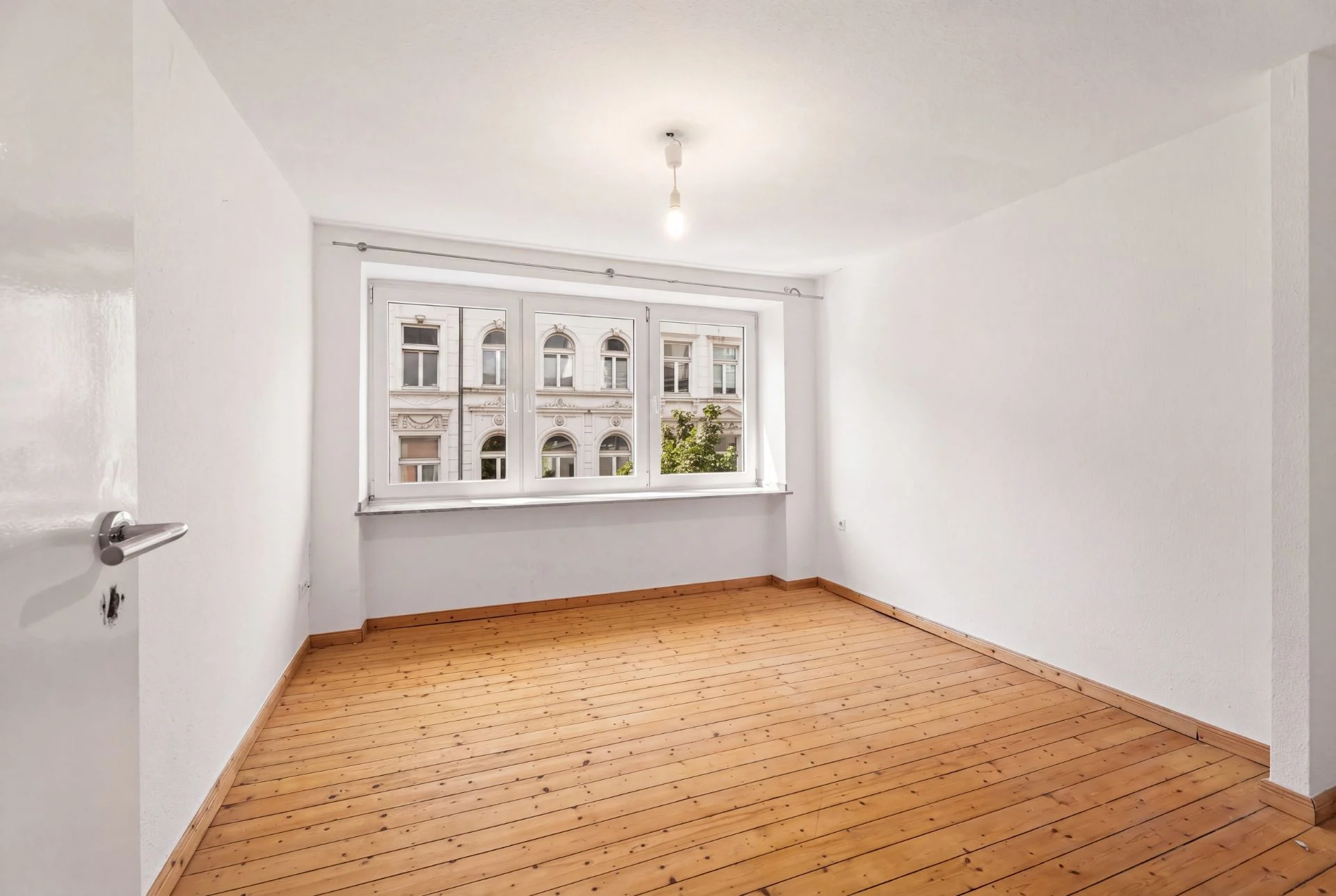 Leerer, heller Raum mit Holzboden und großem Fenster, durch das man Nachbargebäude sieht.