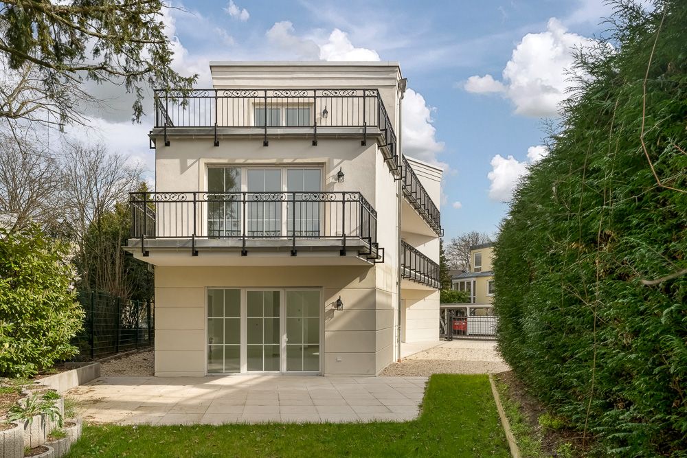 Modernes dreistöckiges Wohnhaus mit mehreren Balkonen und Terrasse, umgeben von Garten und Hecken.