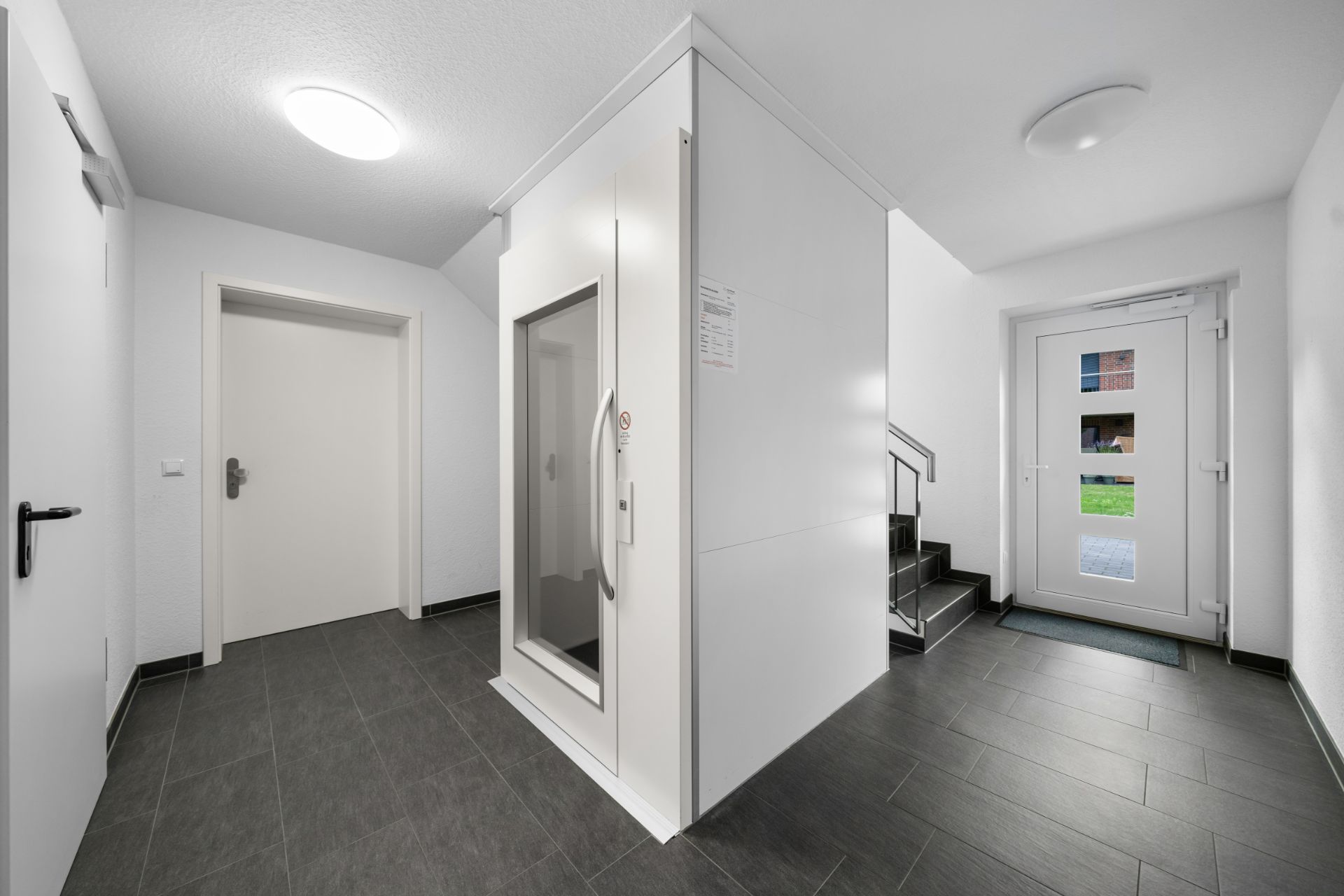 Heller Hausflur mit Aufzug, Treppe und Glastür zum Außenbereich.