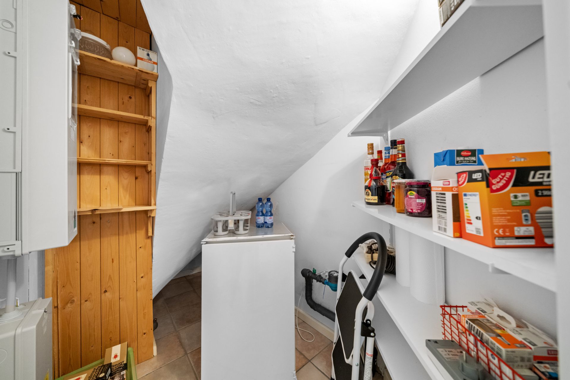 Kleiner Abstellraum mit Regalen, Minikühlschrank und Haushaltsgegenständen unter einer Dachschräge.