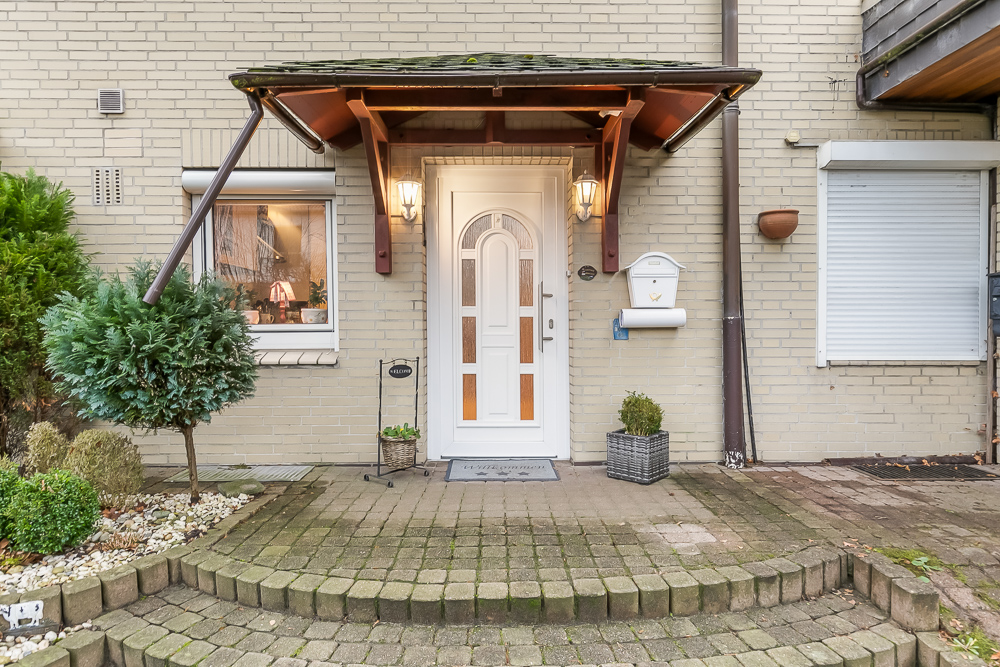 Eingangstür eines Hauses mit Vordach, zwei Lampen, Pflanzen und Briefkasten.