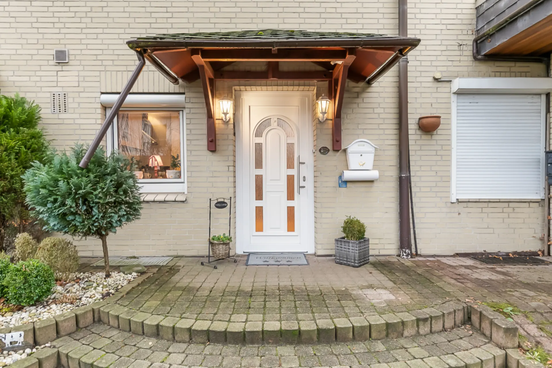 Eingangstür eines Hauses mit Vordach, zwei Lampen, Pflanzen und Briefkasten.