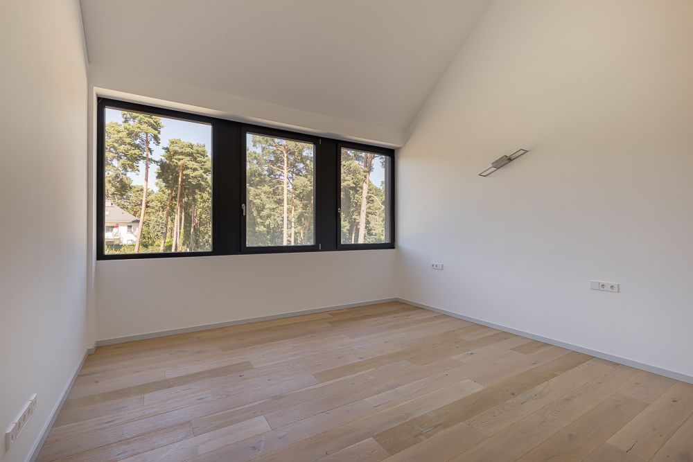 Leeres Zimmer mit Holzfußboden und großem Fenster mit Blick auf Bäume.