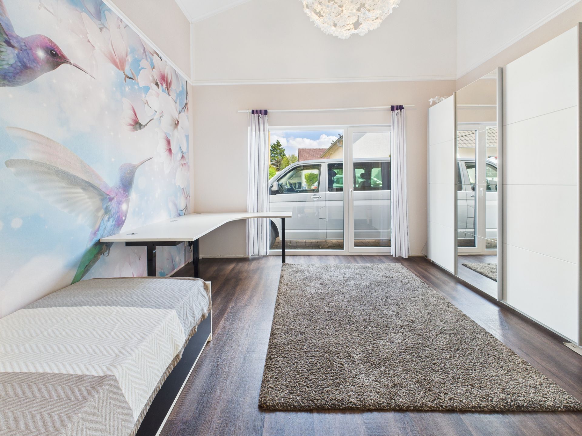 Helles Zimmer mit Teppich, Wandbild mit Kolibri, Bett, Schreibtisch und Spiegel-Schrank.