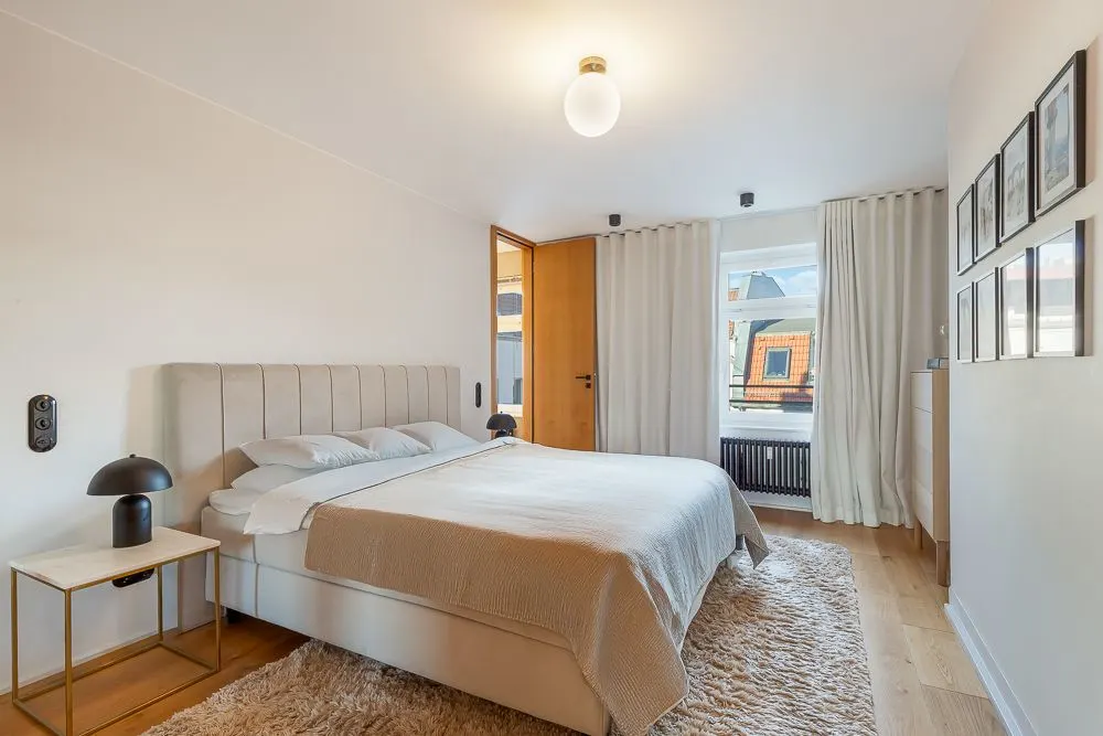 Helles Schlafzimmer mit Doppelbett, Nachttischen, Teppich und Fenstern mit Vorhängen.
