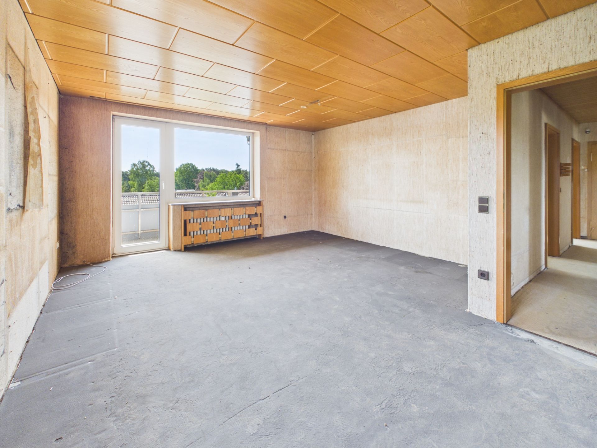 Leerer Raum mit großem Fenster, Holzdecke und rohem Boden, angrenzender Flur sichtbar.