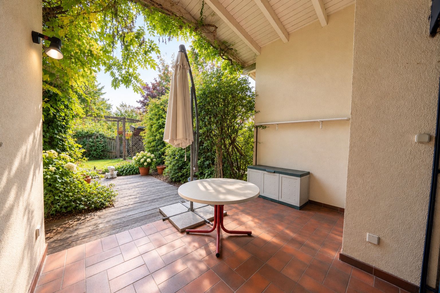 Überdachte Terrasse mit rundem Tisch, Sonnenschirm und Blick in einen Garten.
