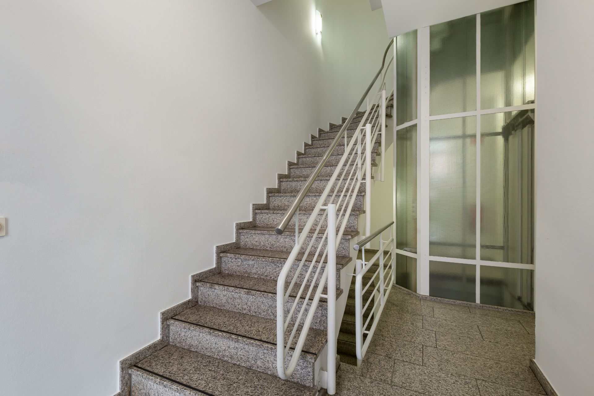 Treppenhaus mit Steintreppe und Geländer, daneben eine Wand mit großen Glasflächen.