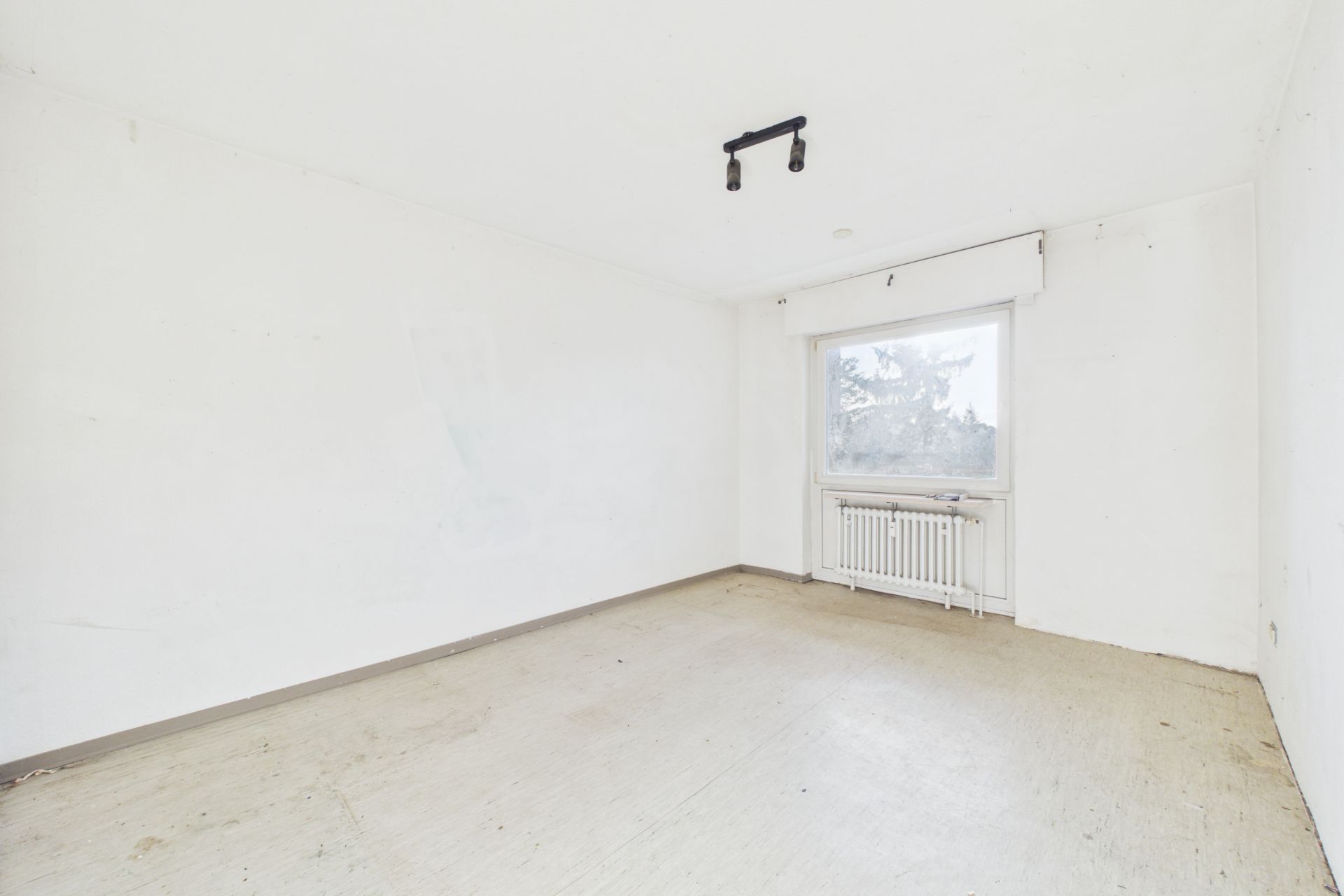 Leerer Raum mit weiß gestrichenen Wänden, großem Fenster und Heizung unter dem Fenster.