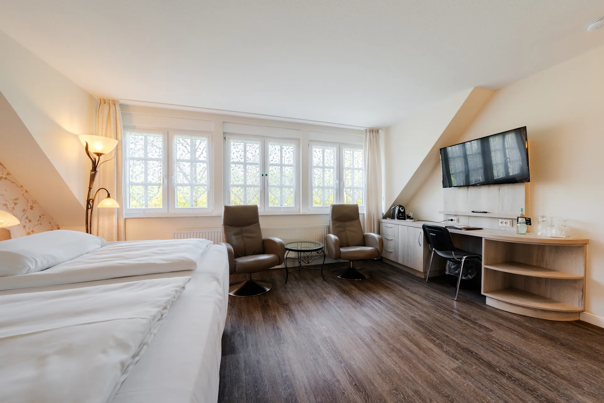 Helles Schlafzimmer mit Doppelbett, zwei Sesseln, Schreibtisch, Fernseher und großen Fenstern.