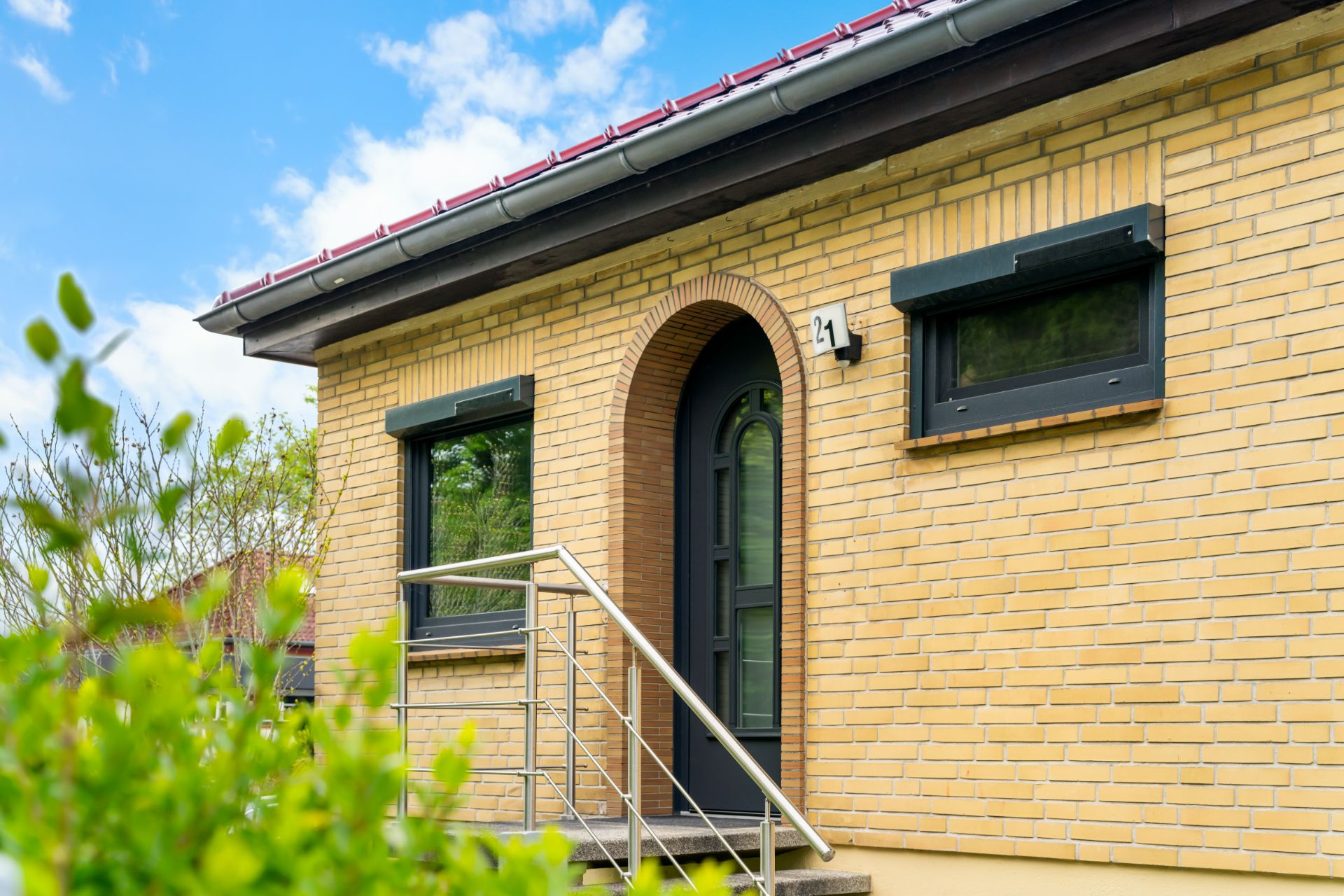 Hausfassade mit gelben Ziegeln, zwei Fenstern und einer Eingangstür mit Treppe.