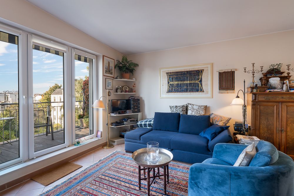 Wohnzimmer mit blauer Couch, Sessel, Teppich und großem Fenster zu einem Balkon.