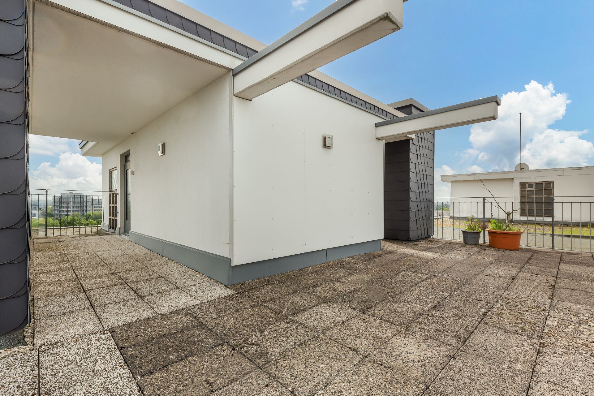 Große Dachterrasse mit Fliesenboden, Geländer und einigen Pflanzkübeln, moderne Hausfassade.