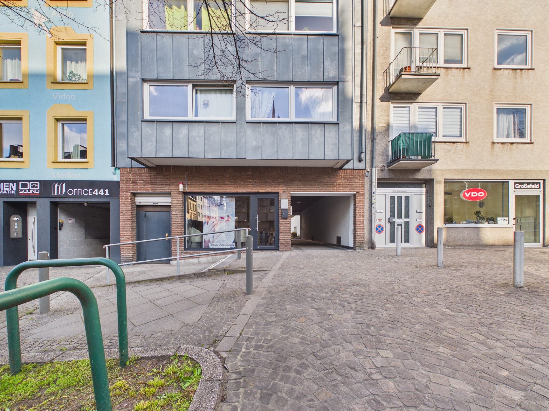 Eingang zu einem Wohnhaus mit Rampe und gepflastertem Vorplatz, flankiert von Geschäften.